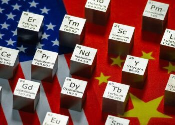 المان‌های مکعبی از عناصر خاکی کمیاب (Rare Earth Elements) که نمادهای شیمیایی مانند Nd، Pr و Er روی آن‌ها حک شده است، و بر روی ترکیبی از پرچم‌های ایالات متحده و چین قرار گرفته‌اند.