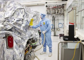 یک تکنسین با لباس محافظ کامل در حال کار بر روی تجهیزات ماشین‌آلات بزرگ ASML که با فویل نقره‌ای پوشانده شده است.