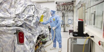 یک تکنسین با لباس محافظ کامل در حال کار بر روی تجهیزات ماشین‌آلات بزرگ ASML که با فویل نقره‌ای پوشانده شده است.