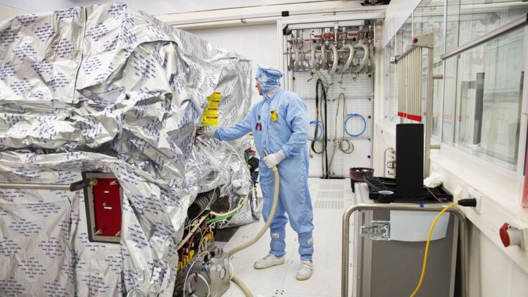 یک تکنسین با لباس محافظ کامل در حال کار بر روی تجهیزات ماشین‌آلات بزرگ ASML که با فویل نقره‌ای پوشانده شده است.