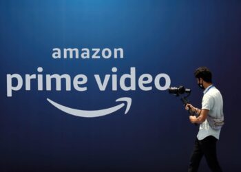 لوگوی Amazon Prime Video به رنگ سفید بر روی یک پس‌زمینه آبی تیره، که یک فرد در حال عبور با تجهیزات دوربین در مقابل آن دیده می‌شود.