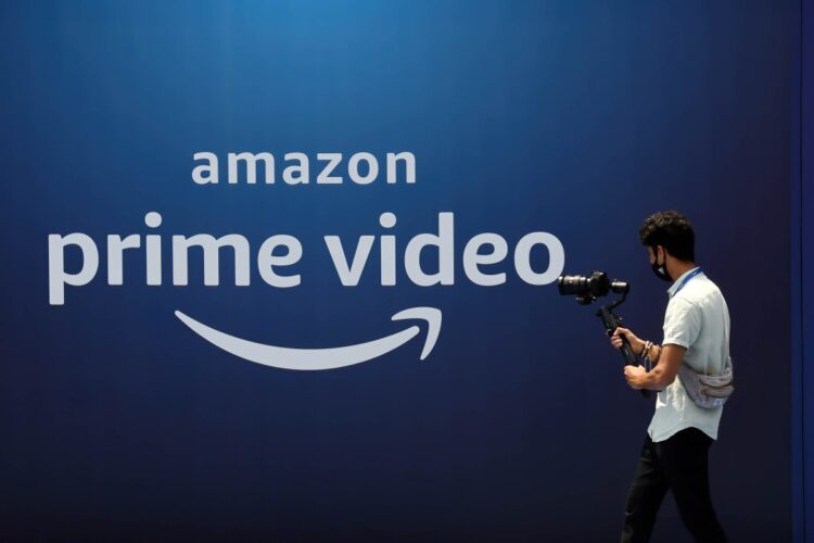 لوگوی Amazon Prime Video به رنگ سفید بر روی یک پسزمینه آبی تیره، که یک فرد در حال عبور با تجهیزات دوربین در مقابل آن دیده میشود.