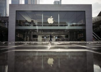 نمای بیرونی یک فروشگاه اپل (Apple Store) با نمای شیشهای بزرگ و لوگوی درخشان اپل در مرکز، که انعکاس آن روی پیادهرو براق دیده میشود.