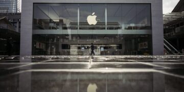 نمای بیرونی یک فروشگاه اپل (Apple Store) با نمای شیشهای بزرگ و لوگوی درخشان اپل در مرکز، که انعکاس آن روی پیادهرو براق دیده میشود.