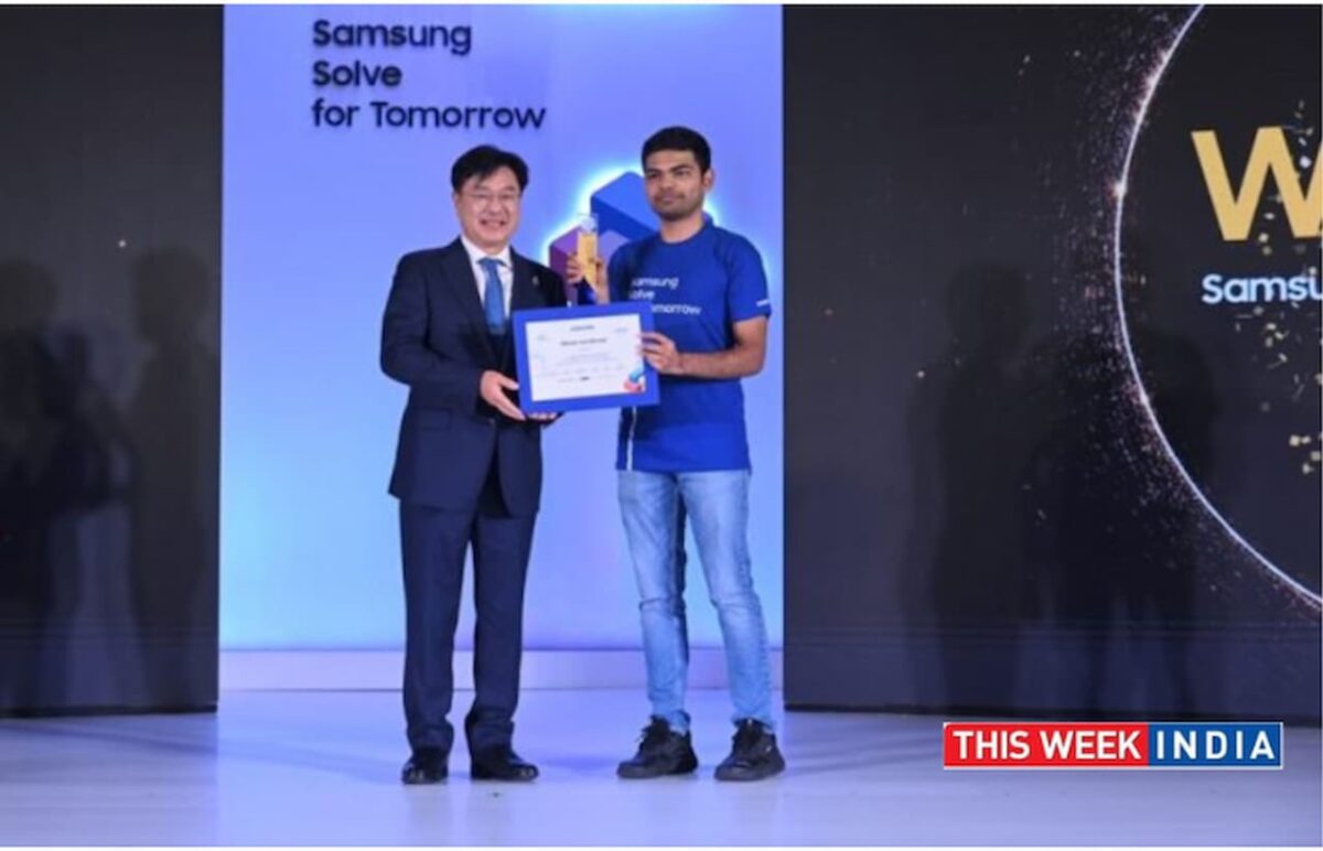 یک مرد کره‌ای با کت و شلوار در حال دادن جایزه (احتمالاً برای مسابقه "Samsung Solve for Tomorrow") به یک دانشجوی هندی که یک گواهی در دست دارد و تی‌شرت "Samsung Solve for Tomorrow" پوشیده است.