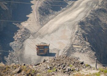 نمای بیرونی معدن روباز بزرگ که کامیون کمپرسی سنگین در حال حمل بار در جاده خاکی پر شیب است.