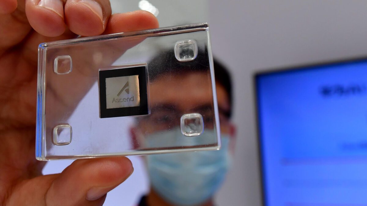 یک دست در حال نگه داشتن یک تراشه با مارک "Ascend" در یک محفظه پلاستیکی شفاف، با انعکاس صورت فردی که ماسک پزشکی زده در پس‌زمینه.