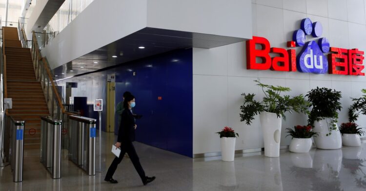 ورودی ساختمان Baidu با پله‌های چوبی، گیت‌های ورودی و لوگوی قرمز و آبی بزرگ Baidu روی دیوار، در حالی که یک کارمند در پیش‌زمینه در حال راه رفتن است.
