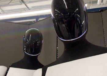 دو ربات انساننما (احتمالاً اپتیموس تسلا) با کلاههای مشکی براق و خطوط نورانی سفید، که از نمای نزدیک در یک فضای داخلی قرار گرفتهاند.