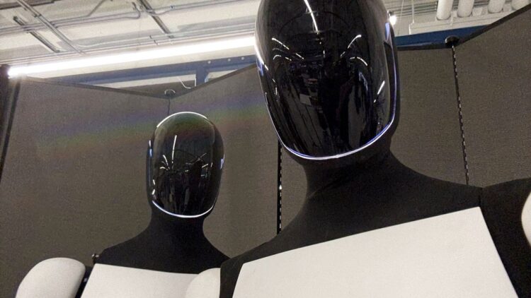 دو ربات انساننما (احتمالاً اپتیموس تسلا) با کلاههای مشکی براق و خطوط نورانی سفید، که از نمای نزدیک در یک فضای داخلی قرار گرفتهاند.