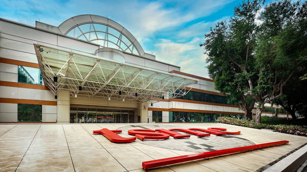نمای بیرونی یک ساختمان TSMC در محوطه سرسبز با لوگوی بزرگ و قرمز شرکت روی زمین.