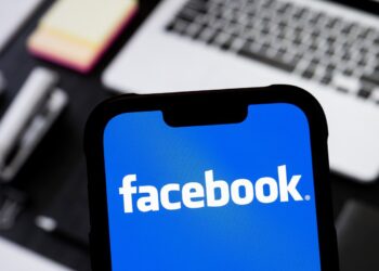 لوگوی سفید رنگ "facebook" بر روی پس‌زمینه آبی روشن، که روی صفحه نمایش یک گوشی هوشمند دیده می‌شود.