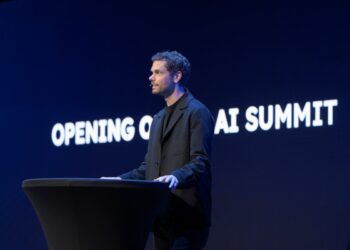 یک مرد در حال سخنرانی در یک کنفرانس (AI Summit) در مقابل یک صفحه نمایش آبی تیره با متن "OPENING OF AI SUMMIT" به صورت برجسته.