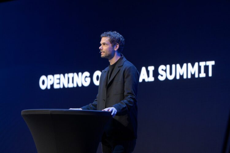 یک مرد در حال سخنرانی در یک کنفرانس (AI Summit) در مقابل یک صفحه نمایش آبی تیره با متن "OPENING OF AI SUMMIT" به صورت برجسته.