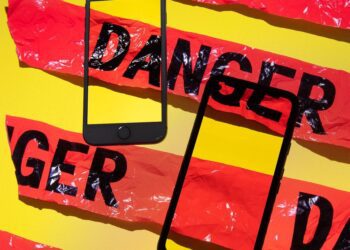 دو گوشی هوشمند مشکی رنگ که به صورت مورب روی پس‌زمینه زرد قرار گرفته‌اند و نوار هشدار پلاستیکی قرمز رنگ با کلمه "DANGER" دور آن‌ها پیچیده شده است.