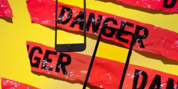 دو گوشی هوشمند مشکی رنگ که به صورت مورب روی پس‌زمینه زرد قرار گرفته‌اند و نوار هشدار پلاستیکی قرمز رنگ با کلمه "DANGER" دور آن‌ها پیچیده شده است.