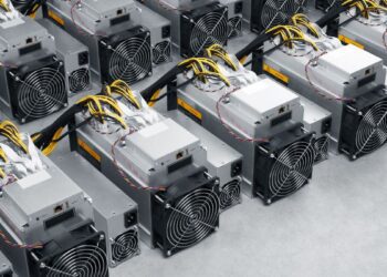 ردیف‌هایی از دستگاه‌های ماینینگ بیت‌کوین (Bitcoin miners) Bitmain Antminer با سیم‌کشی‌های زرد و سیاه که به صورت فشرده روی یک سطح خاکستری چیده شده‌اند.