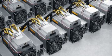 ردیفهایی از دستگاههای ماینینگ بیتکوین (Bitcoin miners) Bitmain Antminer با سیمکشیهای زرد و سیاه که به صورت فشرده روی یک سطح خاکستری چیده شدهاند.