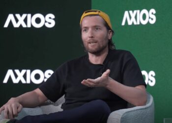الکس کارپ (Alex Karp)، مدیرعامل پالتیر (Palantir)، با کلاه زرد و تی‌شرت مشکی روی صندلی در حال صحبت کردن در یک رویداد Axios.