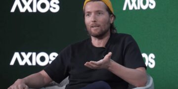 الکس کارپ (Alex Karp)، مدیرعامل پالتیر (Palantir)، با کلاه زرد و تی‌شرت مشکی روی صندلی در حال صحبت کردن در یک رویداد Axios.