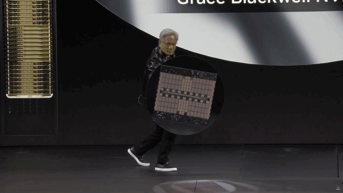 جنسن هوانگ، مدیرعامل انویدیا، در حال قدم زدن روی صحنه و حمل ویفر بزرگ تراشه Blackwell، با پس‌زمینه‌ای تاریک و عناصر طلایی در سمت چپ.