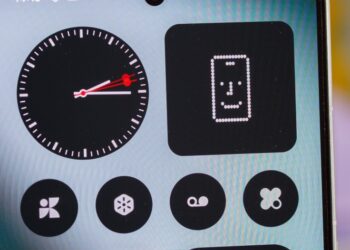 نمای نزدیک از یک گوشی هوشمند (احتمالاً Nothing Phone) که ویجت‌های ساعت آنالوگ و طرحی پیکسلی را در یک محیط تیره و مینیمال نشان می‌دهد.