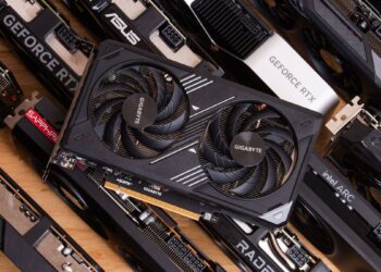 یک کارت گرافیک Gigabyte با دو فن در مرکز، که در میان مجموعه‌ای از کارت‌های گرافیک دیگر با برندهای مختلف (مانند ASUS، SAPPHIRE، Intel ARC) روی یک میز چوبی قرار گرفته است.