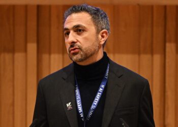 مصطفی سلیمان در حال سخنرانی با یک یقه اسکی مشکی و کت رسمی، با روبان "AI Safety Summit" و پس‌زمینه‌ای از پنل‌های چوبی.