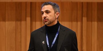 مصطفی سلیمان در حال سخنرانی با یک یقه اسکی مشکی و کت رسمی، با روبان "AI Safety Summit" و پس‌زمینه‌ای از پنل‌های چوبی.