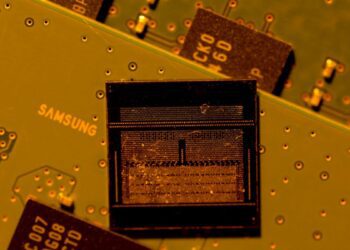 نمای نزدیک از یک تراشه حافظه با بافت داخلی قابل مشاهده، که روی یک برد مدار چاپی (PCB) زرد/زیتونی قرار دارد و لوگوی "SAMSUNG" روی برد دیده می‌شود.