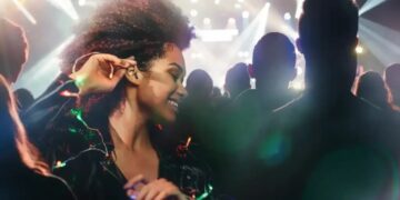 یک زن با موهای فر و لباس براق در حال لبخند زدن و لذت بردن از یک کنسرت یا رویداد موسیقی شلوغ، که دست خود را به گوشش نزدیک کرده است.