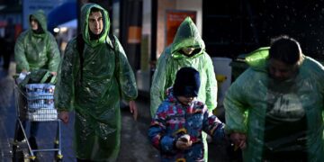 گروهی از افراد در باران، از جمله یک مرد جوان و یک کودک با کلاه و ژاکت پف‌دار، در حال راه رفتن و پوشیدن بارانی‌های پلاستیکی سبز.