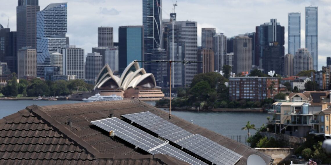 نمایی از بام خانه‌ای با پنل‌های خورشیدی در سیدنی، با پس‌زمینه‌ای از ساختمان اپرای معروف و برج‌های مرکز شهر.