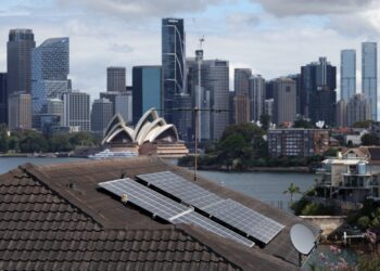 نمایی از بام خانه‌ای با پنل‌های خورشیدی در سیدنی، با پس‌زمینه‌ای از ساختمان اپرای معروف و برج‌های مرکز شهر.