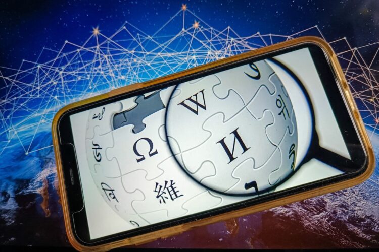 نمای نزدیک از یک گوشی هوشمند که لوگوی ویکی‌پدیا را در یک ذره‌بین روی صفحه نمایش خود نشان می‌دهد، در مقابل یک پس‌زمینه شبکه‌ای آبی.