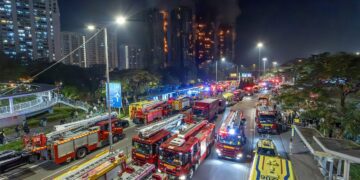 صحنه‌ای در شب از یک آتش‌سوزی بزرگ در طبقات بالایی چندین ساختمان بلند در هنگ کنگ، با تعداد زیادی کامیون آتش‌نشانی و وسایل نقلیه اضطراری در خیابان.