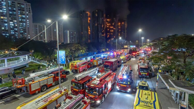 صحنهای در شب از یک آتشسوزی بزرگ در طبقات بالایی چندین ساختمان بلند در هنگ کنگ، با تعداد زیادی کامیون آتشنشانی و وسایل نقلیه اضطراری در خیابان.