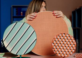 یک زن که در حال نگه داشتن سه بلندگوی دایرهای IKEA با طرحهای مختلف (بافت نارنجی، نوارهای سبز و الگوی هندسی) در یک محیط داخلی رنگارنگ است.