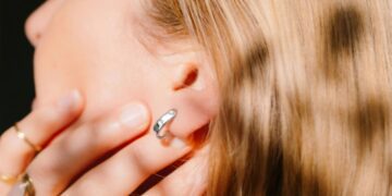 نمای نزدیک از یک زن با موهای بلوند که یک حلقه نقرهای یا حسگر پوشیدنی در لاله گوشش دارد و با دست روی صورت خود را پوشانده است.