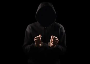 نمای سیلوئت یک فرد با هودی تیره که دستبند فلزی به دستان دارد، در پسزمینه کاملاً مشکی، تصویر مفهومی از جرم سایبری، بازداشت یا فعالیت غیرقانونی دیجیتال