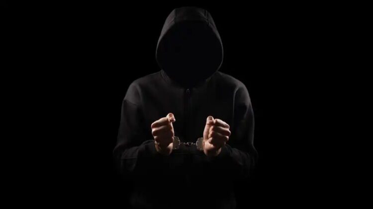 نمای سیلوئت یک فرد با هودی تیره که دست‌بند فلزی به دستان دارد، در پس‌زمینه کاملاً مشکی، تصویر مفهومی از جرم سایبری، بازداشت یا فعالیت غیرقانونی دیجیتال