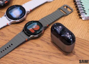 دو عدد ساعت هوشمند گلکسی واچ با بندهای سفید و زیتونی در کنار کیس هدفون گلکسی بادز روی میز.