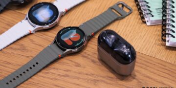 دو عدد ساعت هوشمند گلکسی واچ با بندهای سفید و زیتونی در کنار کیس هدفون گلکسی بادز روی میز.