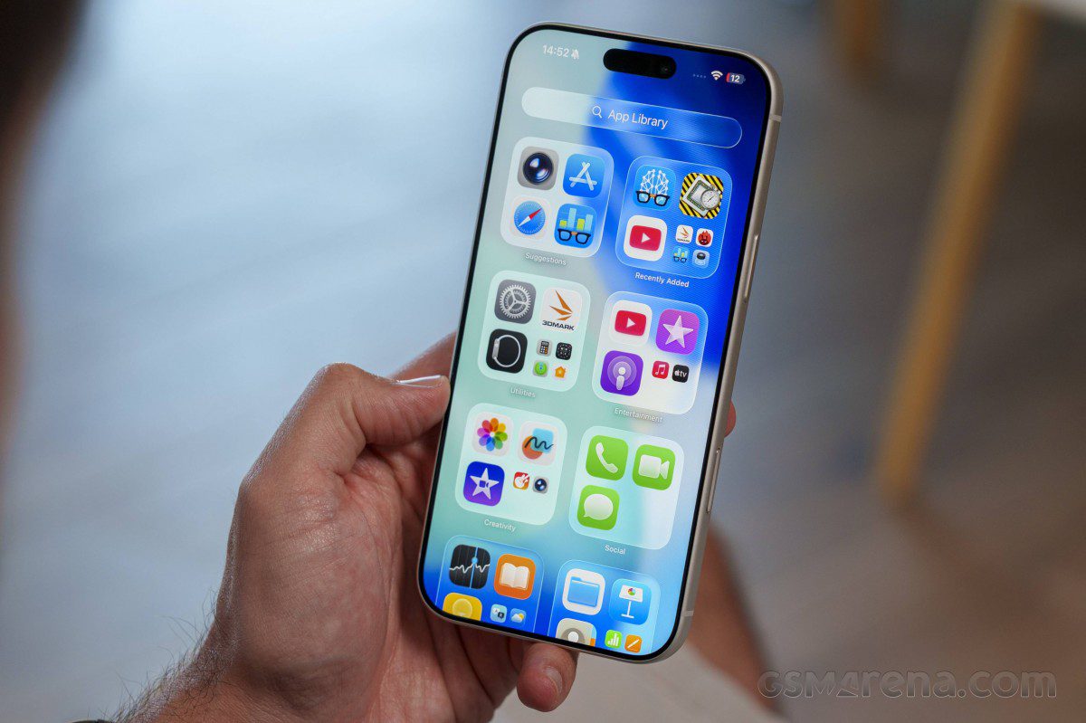 نمای نزدیک از یک گوشی آیفون در دست که صفحه «App Library» و آیکون برنامه‌های مختلف را نشان می‌دهد.