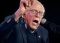 برنی سندرز پشت میکروفون در حال سخنرانی با حالت چهره‌ای هیجان‌زده و انگشت اشاره رو به بالا.