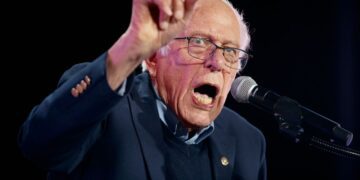 برنی سندرز پشت میکروفون در حال سخنرانی با حالت چهره‌ای هیجان‌زده و انگشت اشاره رو به بالا.