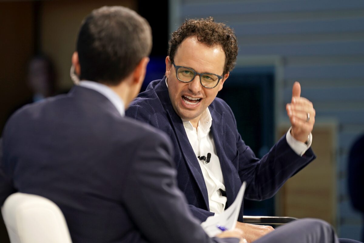 مردی با موهای فر و عینک که کت‌وشلوار سرمه‌ای پوشیده و در حال صحبت و ژست گرفتن با دست در یک نشست است.