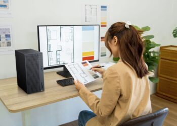 زنی در حال کار با کامپیوتر ایسوس ExpertCenter و بررسی نقشههای معماری روی میز کار.