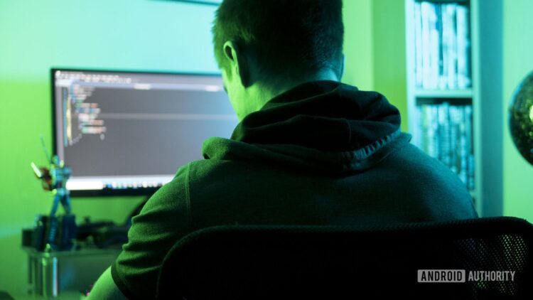 تصویر نمادین از یک هکر با هودی که در اتاقی با نور سبز جلوی کامپیوتر نشسته است.