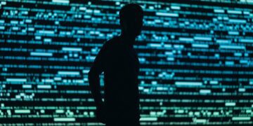 شبح یک مرد در مقابل یک صفحه نمایش دیجیتالی روشن، متشکل از خطوط کد آبی و سبز، نمادی از دادهها و امنیت.